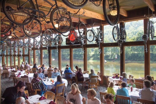 A restaurant with hanging bicycles, filled with diners, overlooking a scenic river view through large windows.