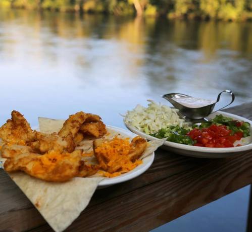 A plate of fried fish with cheese on tortillas, accompanied by chopped vegetables and a sauce, by a river.