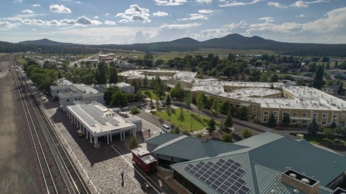Aerial view of a small town with buildings, trees, and mountains in the background under a partly cloudy sky.