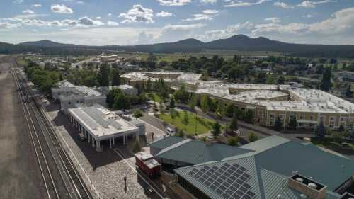 Aerial view of a small town with buildings, trees, and mountains in the background under a partly cloudy sky.