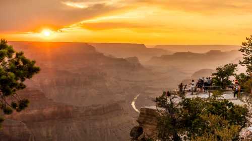 Sunset over the Grand Canyon, with visitors enjoying the view from a scenic overlook surrounded by trees.