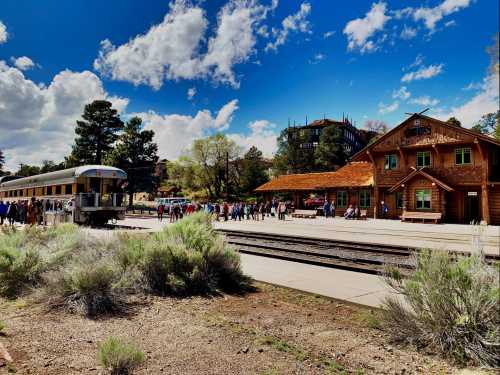 A train station with a vintage train, surrounded by people and trees, under a bright blue sky with fluffy clouds.