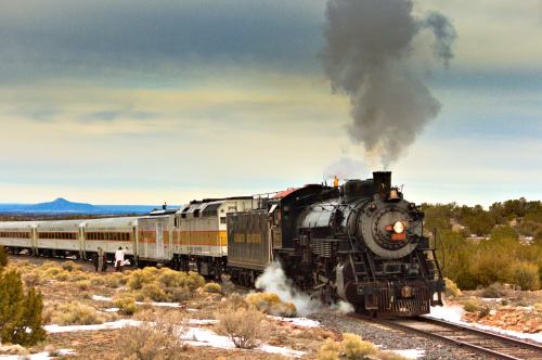 A vintage steam train billows smoke as it travels through a desert landscape under a cloudy sky.