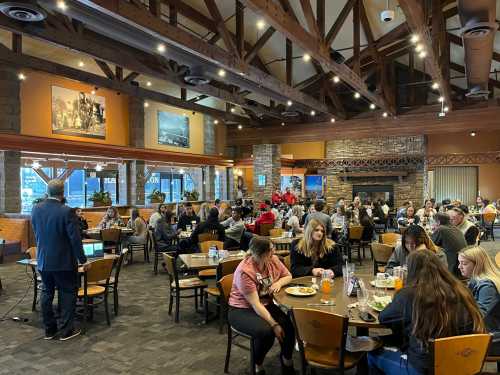 A busy restaurant interior with wooden beams, filled with diners enjoying meals and a speaker at the front.