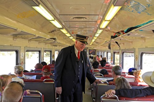 A train conductor in uniform interacts with passengers inside a vintage train car filled with seated travelers.