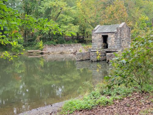 A small stone building by a calm pond, surrounded by trees with autumn foliage reflecting in the water.