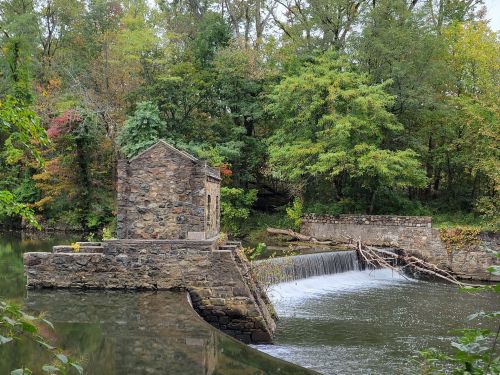 A stone structure beside a river, featuring a small waterfall and surrounded by lush green trees in autumn colors.