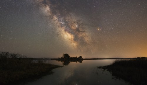 A serene night sky filled with stars and the Milky Way, reflecting on a calm water surface near a silhouette of trees.