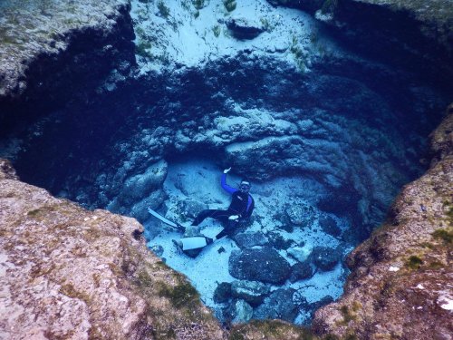 A diver sits at the bottom of a deep underwater sinkhole, surrounded by rocky walls and clear blue water.