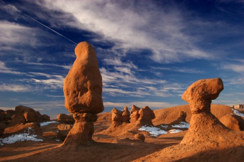 Unique rock formations rise from a sandy landscape under a blue sky with wispy clouds. Snow patches are visible.