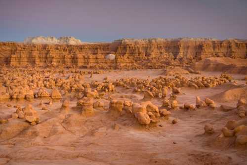 A vast desert landscape with unique rock formations and layered cliffs under a soft, colorful sky.