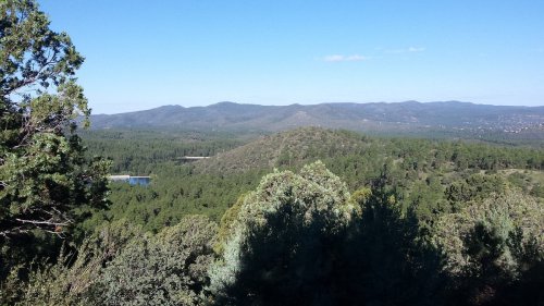 A panoramic view of lush green mountains under a clear blue sky, with a hint of a lake in the distance.