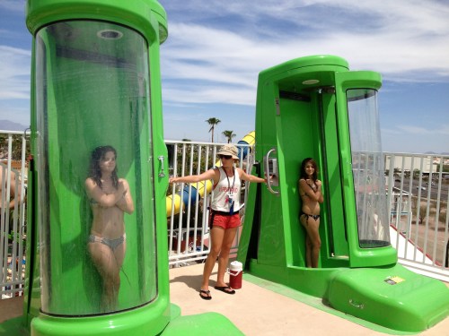 Two women in bikinis stand in green shower-like structures, while a lifeguard supervises nearby on a sunny day.