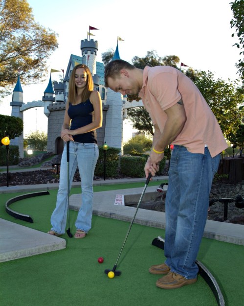 A man and woman play mini-golf on a green course with a castle in the background during sunset.