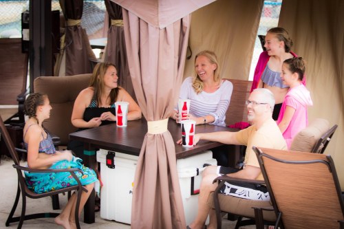 A group of six people, including children and adults, laughing and enjoying drinks around a table in a shaded outdoor area.