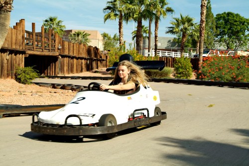 A young girl with long hair races in a go-kart on a sunny track surrounded by palm trees and greenery.