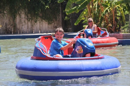 A smiling boy in a blue bumper boat, enjoying a sunny day on the water, with another boat and a woman in the background.