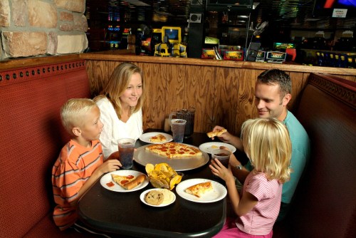 A family of four enjoys pizza and snacks at a restaurant, seated at a booth with drinks and plates of food.