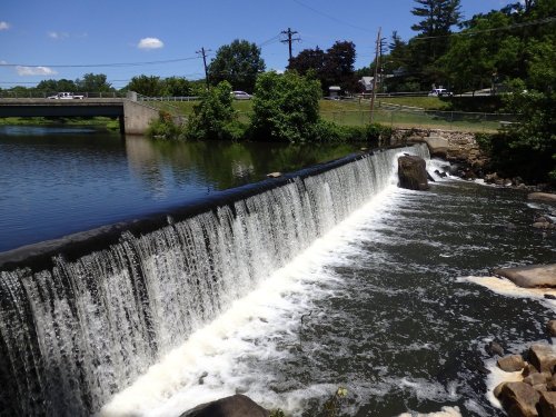 A waterfall cascades over a dam into a calm river, surrounded by greenery and a clear blue sky.