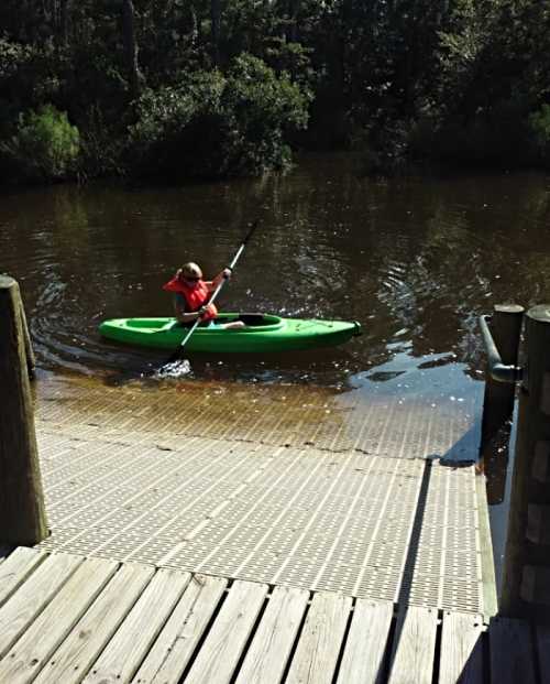 A person in a green kayak paddles on a calm river, with a wooden dock in the foreground and trees in the background.