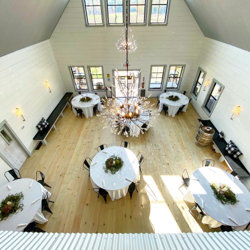A spacious event room with round tables, elegant decor, and a large chandelier, featuring wooden floors and bright windows.