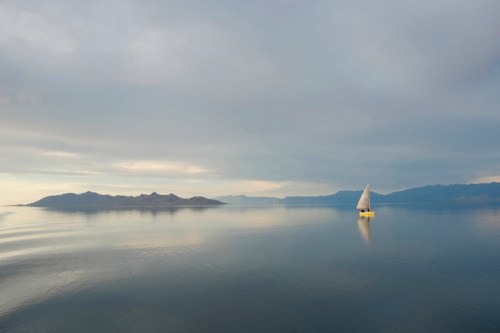 A solitary sailboat glides across calm waters, with distant mountains under a cloudy sky.
