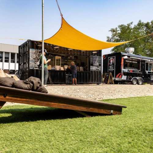 A food truck setup with a shaded area, people ordering, and a grassy space in front under a clear blue sky.