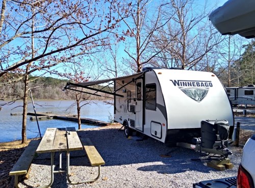 A Winnebago camper parked by a lake, surrounded by trees and a picnic table nearby. Clear blue sky above.