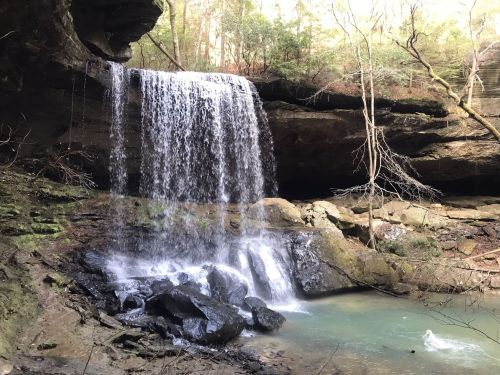 A serene waterfall cascades over rocks into a tranquil pool, surrounded by lush greenery and rugged cliffs.