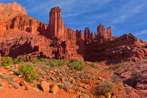 Red rock formations rise against a blue sky, showcasing rugged cliffs and scattered greenery in a desert landscape.