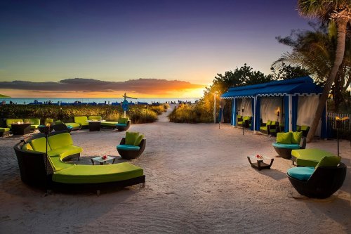 Lounge area with green seating on a beach at sunset, with palm trees and a cabana in the background.