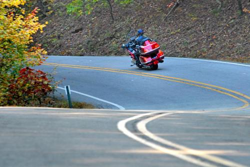 A motorcyclist rides a red bike along a winding road surrounded by autumn foliage.