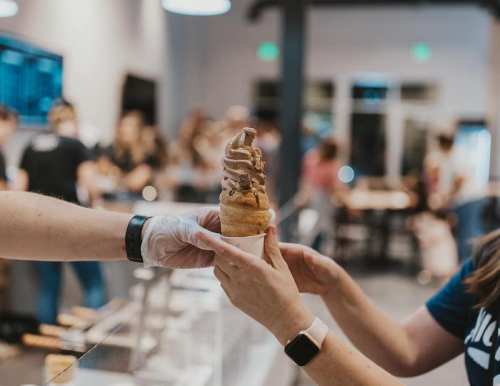 A hand in a glove holds a soft-serve ice cream cone, while another hand reaches to take it in a busy ice cream shop.
