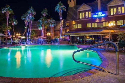 A brightly lit pool at night, surrounded by palm trees and a hotel with a glowing sign in the background.