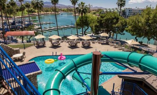 A vibrant pool area with a water slide, lounge chairs, and a scenic view of a lake and palm trees.