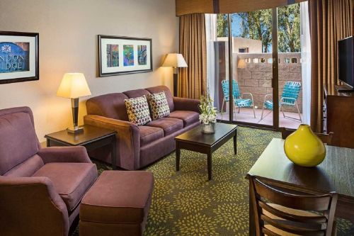 Cozy hotel room with a sofa, armchair, coffee table, and patio access, featuring warm lighting and colorful decor.