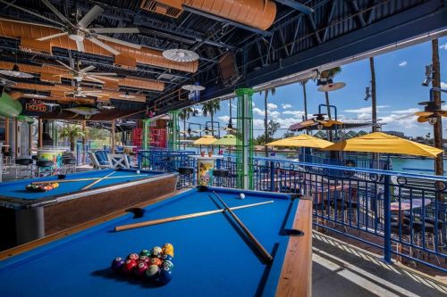 A vibrant outdoor bar area with a pool table, colorful umbrellas, and palm trees under a clear blue sky.
