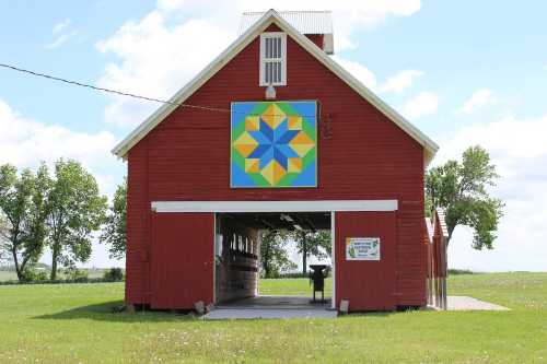 A red barn with a colorful quilt pattern on the front, set against a blue sky and green fields.