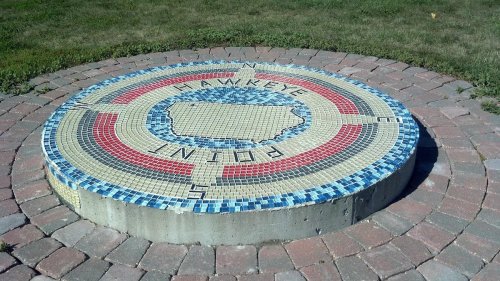 A circular mosaic featuring a map outline and directional markers, set in a grassy area with stone paving.