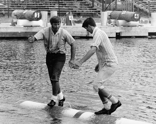 Two boys balancing on a log over water, one reaching out to help the other, in a playful outdoor setting.