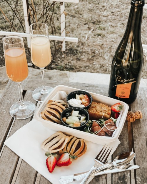 A picnic spread featuring pastries, fruit, and a bottle of Prosecco, with two glasses of sparkling drinks on a wooden table.