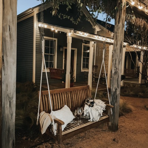 A cozy outdoor swing bench with blankets and wine glasses, illuminated by string lights near a charming house.