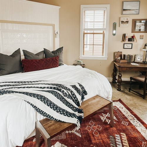 Cozy bedroom with a bed, decorative pillows, a wooden table, and a patterned rug. Natural light from a window.
