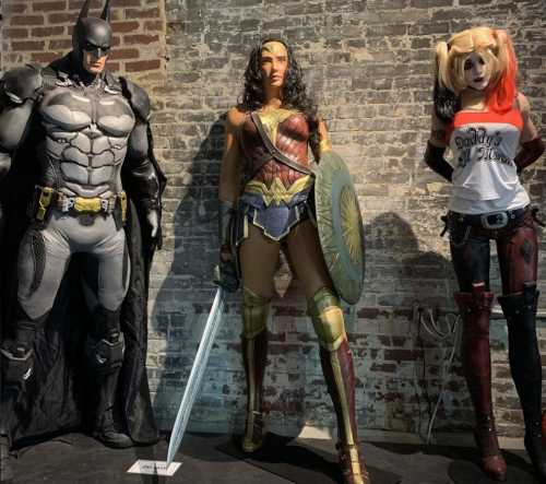 Three life-sized superhero figures: Batman, Wonder Woman, and Harley Quinn, displayed against a brick wall.