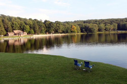 Two blue chairs sit on a grassy shore by a calm lake, surrounded by trees and a distant building.