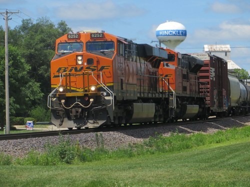 A BNSF freight train with two locomotives passes by a water tower in Hinckley, surrounded by green grass and trees.
