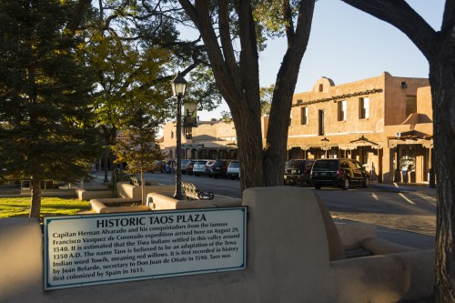 Historic Taos Plaza with adobe buildings, trees, and a sign detailing its history and significance.