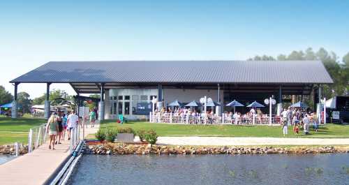 A waterfront venue with a large patio, people dining outside, and a dock leading to the building.