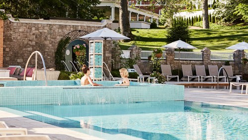 Two people relax in a turquoise pool surrounded by lounge chairs and umbrellas in a serene outdoor setting.