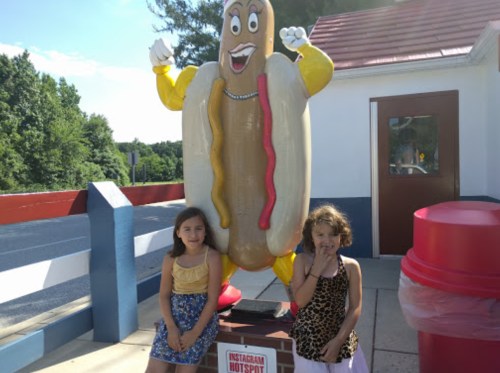 Two girls stand beside a large, smiling hot dog statue at a roadside location, surrounded by greenery.